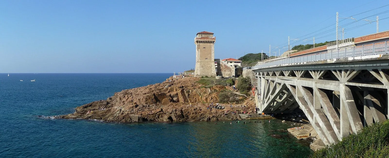 La Torre di Calafuria su un promontorio di roccia rossastra a picco sul mare blu, con bagnanti sugli scogli e in acqua e, a destra, il ponte in cemento della strada e della ferrovia