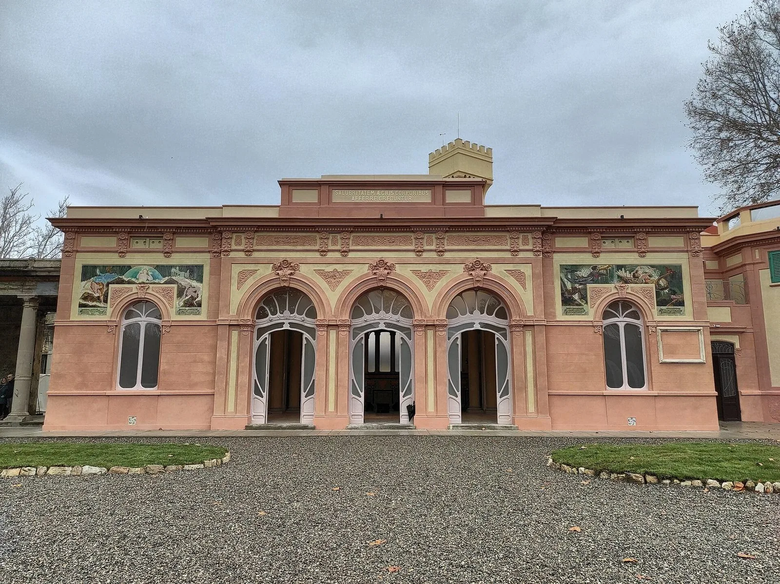 La facciata restaurata della Sala della Mescita alle Terme del Corallo di Livorno, rosa e ocra, con tre grandi arcate liberty, pannelli dipinti e un vialetto di ghiaia