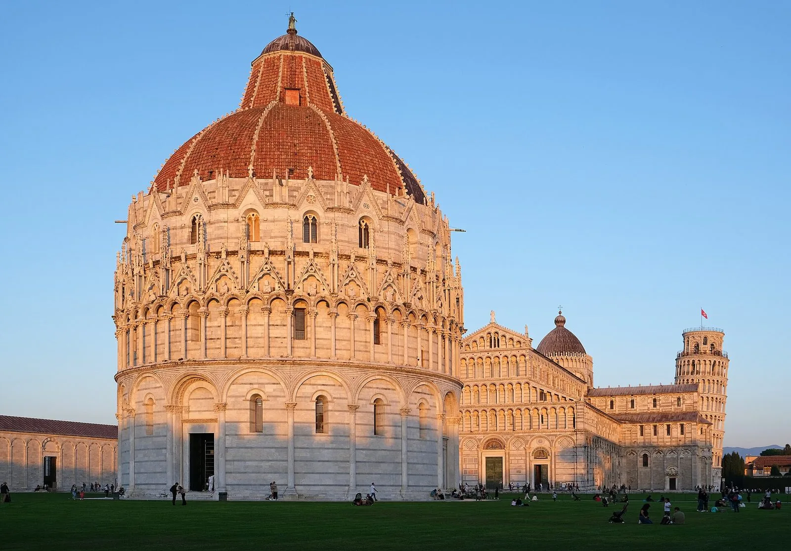 Il Battistero di Pisa illuminato dal sole del tardo pomeriggio, con il Duomo e la Torre pendente sullo sfondo e il prato della Piazza dei Miracoli